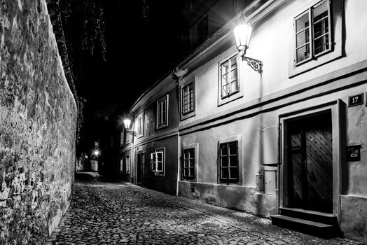 Narrow cobbled street in old medieval town with illuminated houses by vintage street lamps, Novy svet, Prague, Czech Republic. Night shot. Black and white image.