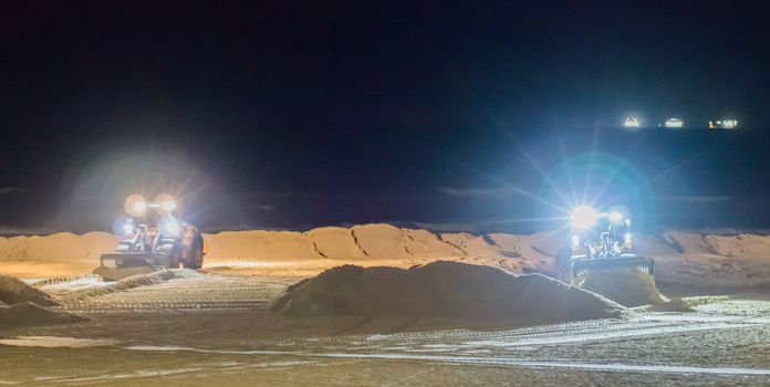 two ground workers working at night in bulldozers at the beach moving the sand