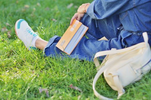 Beautiful, happy young student girl sitting on green grass near the backpack, bag under the tree near the campus, university, school, education. Summer, spring green park