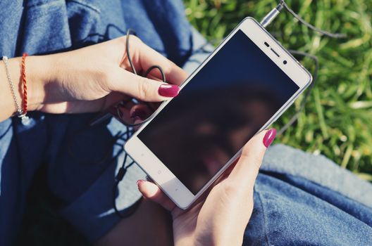 Hands of a young girl who holds a smartphone and sits on the green grass, close-up