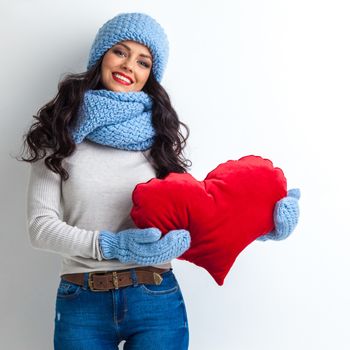 Beautiful smiling brunette woman in winter hat and scarf holding a red heart pillow