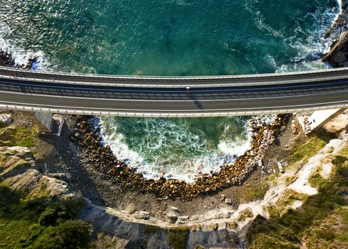 High above near the cliffs views down onto the Sea Cliff Bridge as a cyclist crosses leaving long shadows in the early morning light