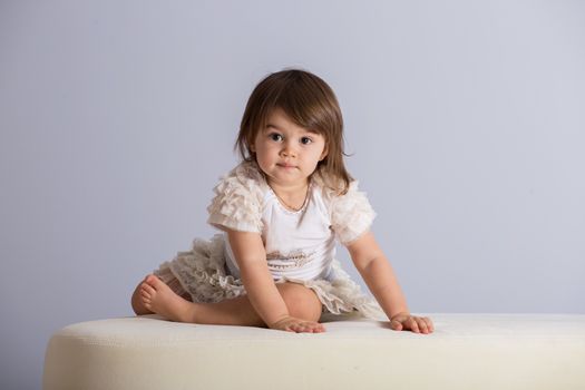 Pretty baby girl with brown hair in white dress on light purple background