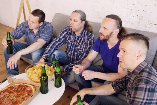 Group of men drinking beer, eating pizza, talking and smiling while resting at home on couch behind TV