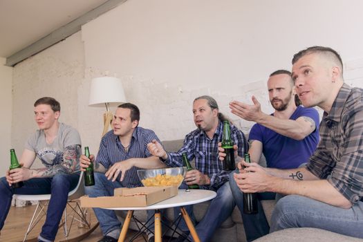 Male friends sports fans watching loosing football match on TV at home on couch sharing snacks drinking beer