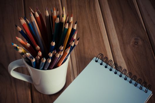 Color pencils in the cups and notebook placed on a desk.