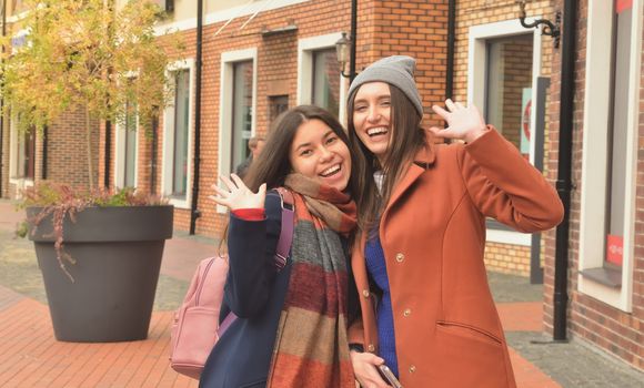 Two girlfriends in coats walking around the city, smiling and waving at the camera