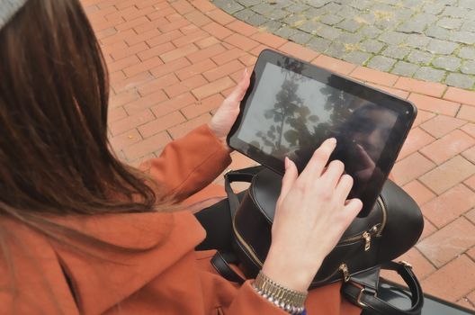 The girl in the coat sit on the bench and uses the tablet