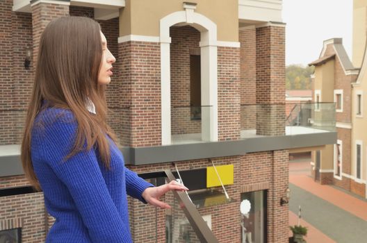 A beautiful slim girl is standing on the balcony of a shopping center, looking at the building