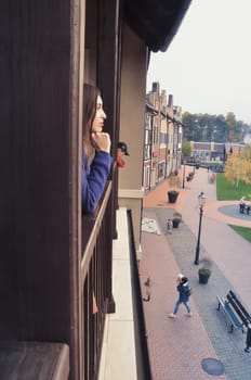 The European girl leans on the window sill and looks at the trees and buildings