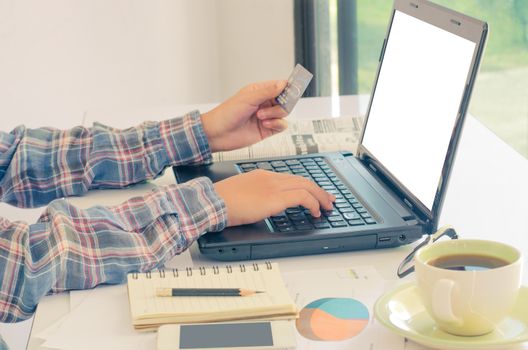View of male hands holding credit card numbers typed on the computer keyboard while sitting at a desk - focus hand.