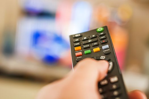 A hand pressing a remote control button to change channels in front of a blurry television screen