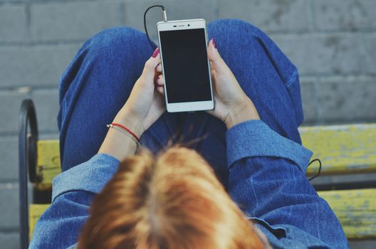 A young pretty student uses a smartphone sitting on a yellow bench in a denim jacket on the street