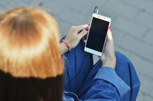 Smartphone close-up in the hands of a girl dressed in denim clothing that listens to music in headphones, side view