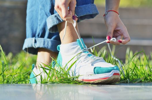 Young girl wearing jeans tying shoelaces on sneakers standing on green grass side view close-up horizontal photo, Sunny day