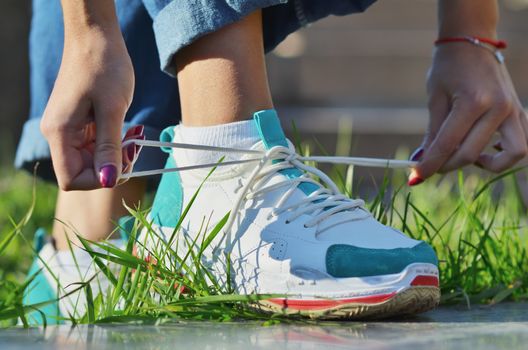 Young girl wearing jeans tying shoelaces on sneakers standing on green grass side view close-up horizontal photo, Sunny day