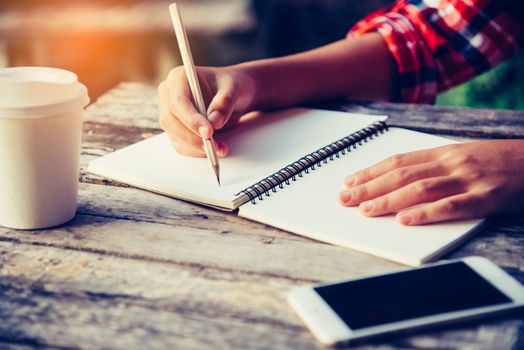 The hand is writing a notebook with a pencil and a cup of coffee, a smartphone placed on a wooden desk in an office with golden light.
