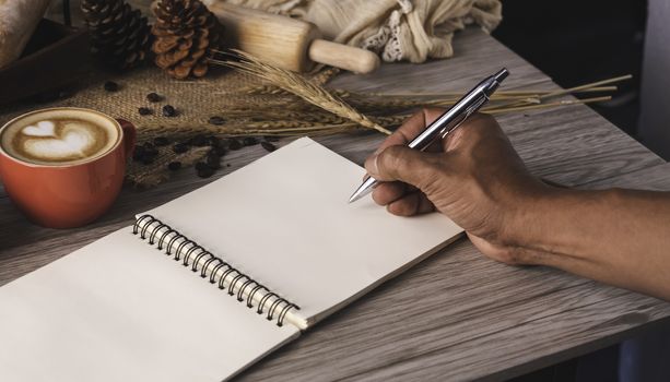 Hand holding pen is writting white notebook spread on a table with coffee and bread placed.