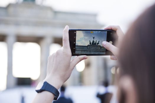 Hands holding phone mobile and takes pictures of famous historical tourist sites. The Brandenburg Gate neoclassical monument in Berlin.