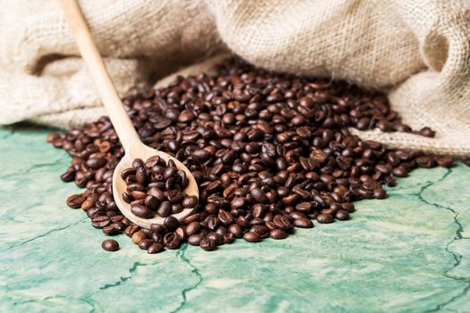 Coffee beans in coffee burlap bag on green surface and wooden spoon with coffee beans on top.