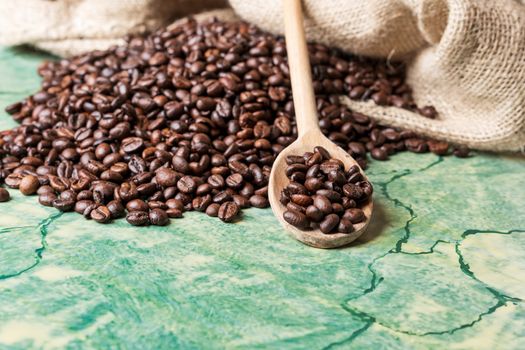 Coffee beans in coffee burlap bag on green surface and wooden spoon with coffee beans on top.