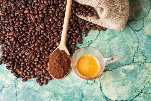 Coffee beans in coffee burlap bag on green surface,wooden spoon with ground coffee on top and coffee glass cup, view from above