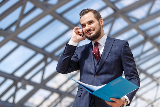 Portrait of mature businessman holding binder and documents and talking by phone over modern building interior background