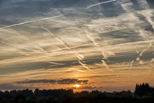 sunset in august in the belgium ardennes with clouds and lot of stripes from the planes in the sky and the red orange sun going under