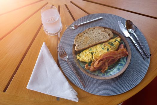 Bread, omelette and bacon in a plate with spoon knife and fork with a glass of water on the wooden table. The light shining from top.