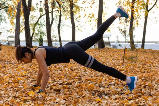 Attractive woman in sports clothes doing sports exercises in nature, on the carpet of autumn leaves, loves gymnastics, kneads his legs. Active young girl engaged in sports, leads a healthy lifestyle.