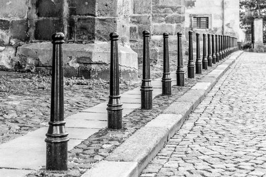 Row of iron pillars between cobbled street and pedestrian sidewalks. Black and white image.