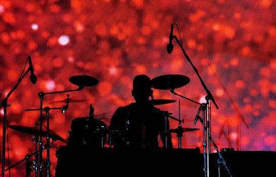 A silhouette of a drummer in a music concert against a red pattern screen behind.                               