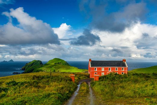 A red house in the Ireland countryside. The cloudy sky contrasting with the green of the fields and the sea is vissible in the background. Vivid colours and great contrasts.