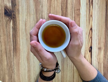 A closeup of a woman's hands holding a cup of hot tea. The background is wooden table. One of the bracelets is made of leather and the other is silver