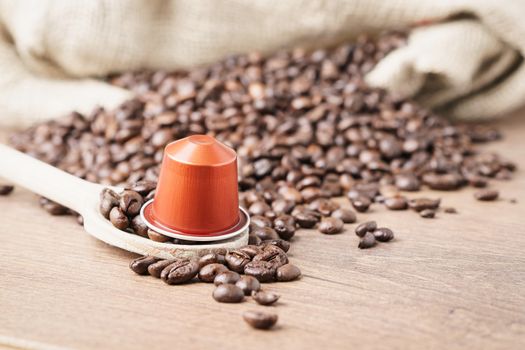 In the foreground a coffee capsule on wooden spoon and   roasted coffee beans with burlap sack on blur wooden background ,close up.