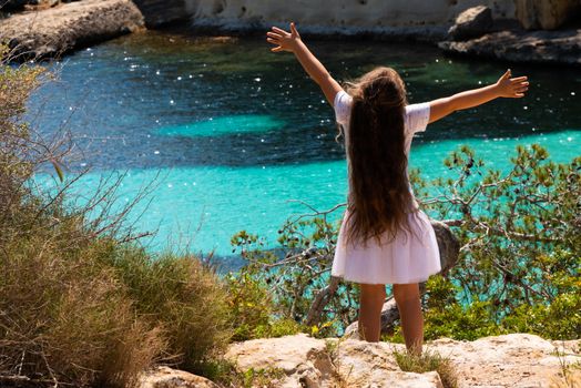 Opened arm girl with long hair watching the crystal clear water of Mediterranean sea in Mallorca.