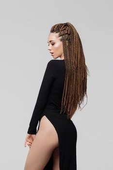 Side view of sensual young female with long dreadlocks wearing amazing black dress and looking at camera while standing on light gray background