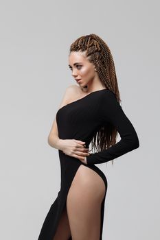 Side view of sensual young female with long dreadlocks wearing amazing black dress and looking at camera while standing on light gray background