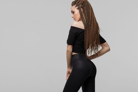 Beautiful young woman with dreadlocks keeping hand on waist and looking at camera while standing on light gray background