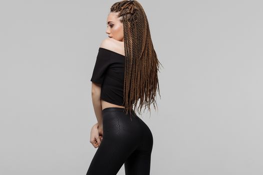 Beautiful young woman with dreadlocks keeping hand on waist and looking at camera while standing on light gray background