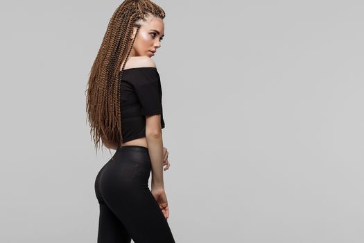 Beautiful young woman with dreadlocks keeping hand on waist and looking at camera while standing on light gray background