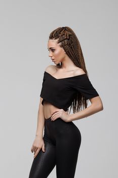 Beautiful young woman with dreadlocks keeping hand on waist and looking at camera while standing on light gray background