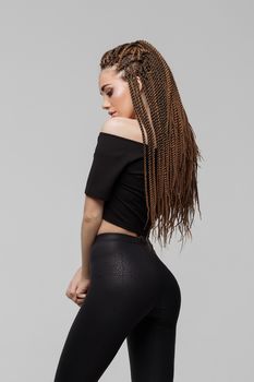 Beautiful young woman with dreadlocks keeping hand on waist and looking at camera while standing on light gray background