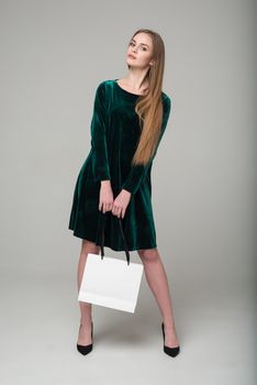 Young model long-haired blond girl in dark green short dress with lace stands holding white package with copy space