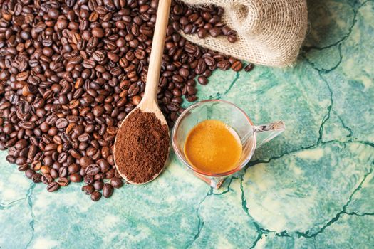 Coffee beans in coffee burlap bag on green surface,wooden spoon with ground coffee on top and coffee glass cup, view from above