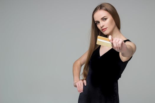 Young model long-haired blond girl in black dress holds golden banking credit card for shopping payment