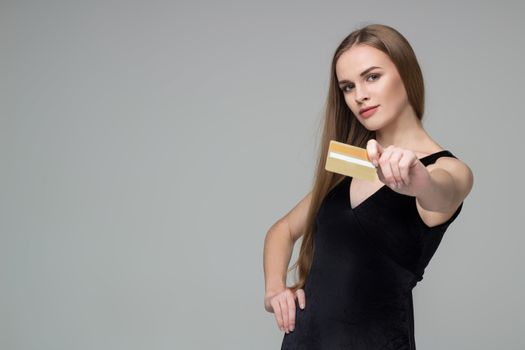 Young model long-haired blond girl in black dress holds golden banking credit card for shopping payment