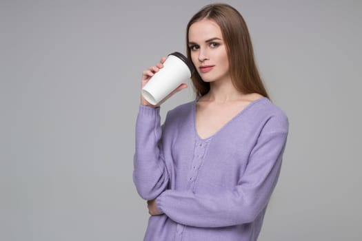 Young model long-haired blond girl in blue purple lilac jamper holds paper plastic coffee glass drinking