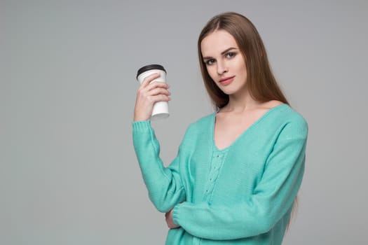 Young model long-haired blond girl in blue aquamarine jamper holds paper plastic coffee glass drinking