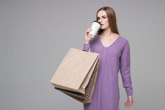 Young model long-haired blond girl in blue purple lilac jamper holds shopping paper bags and  plastic coffee glass drinking